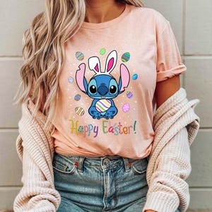 May include: A pink t-shirt with a blue cartoon character wearing bunny ears and holding a colorful Easter egg. The character is surrounded by colorful Easter eggs. The text "Happy Easter!" is printed below the character.
