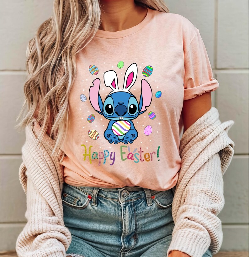 May include: A pink t-shirt with a blue cartoon character wearing bunny ears and holding a colorful Easter egg. The character is surrounded by colorful Easter eggs. The text "Happy Easter!" is printed below the character.