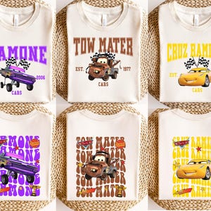 May include: Six off-white t-shirts with different designs featuring characters from the movie Cars. The designs include the names "Ramone", "Tow Mater", and "Cruz Ramirez" with associated graphics and text. The shirts are arranged on a woven surface.