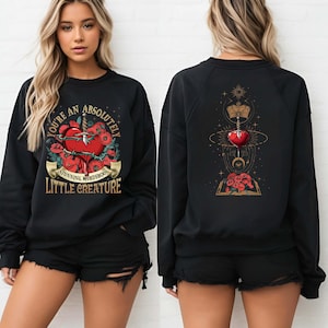 May include: Black sweatshirt with a graphic print of a heart with a dagger through it and the text "You're an absolutely stunning murderous little creature". The back of the sweatshirt has a graphic print of a heart with a dagger through it, a crown, and a book with flowers.