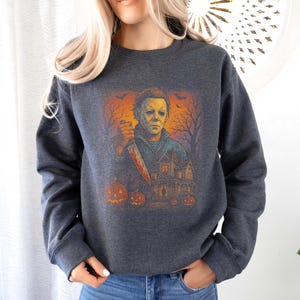 May include: Dark gray sweatshirt featuring a Halloween-themed graphic. The graphic depicts a character holding a bloody knife in front of a haunted house, with pumpkins and a spooky orange sky. The sweatshirt has long sleeves and a crew neck.