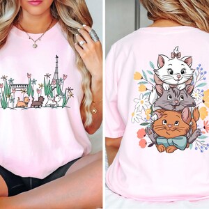 May include: Pink t-shirt with a graphic of three cats from the Disney movie The Aristocats. The cats are stacked on top of each other and surrounded by flowers. The front of the shirt has a graphic of the Eiffel Tower and three cats.