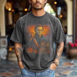 May include: Dark gray t-shirt featuring a Halloween-themed graphic. The graphic depicts a figure holding a bloody knife, a haunted house, pumpkins, and bats against an orange and black background. The shirt has short sleeves.