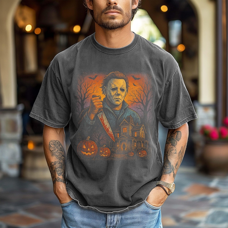 May include: Dark gray t-shirt featuring a Halloween-themed graphic. The graphic depicts a figure holding a bloody knife, a haunted house, pumpkins, and bats against an orange and black background. The shirt has short sleeves.