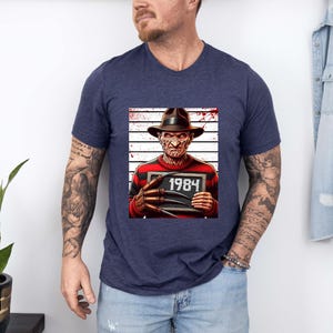 May include: Navy blue v-neck t-shirt featuring a graphic of a horror movie character holding a sign that reads "1984". The character is wearing a hat and a red and black striped sweater. The background has a white and gray striped pattern.