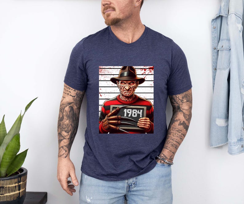 May include: Navy blue v-neck t-shirt featuring a graphic of a horror movie character holding a sign that reads "1984". The character is wearing a hat and a red and black striped sweater. The background has a white and gray striped pattern.