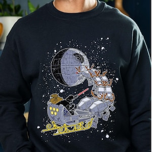 May include: Black sweatshirt featuring a Star Wars-themed holiday design. The graphic depicts Darth Vader in a sleigh pulled by AT-ATs, with the Death Star in the background. A red lightsaber is visible. The design is set against a starry sky.