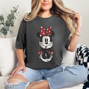 May include: A woman wearing a dark gray t-shirt with a vintage-style image of Minnie Mouse. The t-shirt has a distressed look and features Minnie Mouse with a red bow and white polka dots. The woman is sitting on a white couch with a black and white checkered throw blanket.