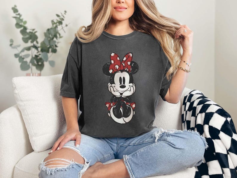 May include: A woman wearing a dark gray t-shirt with a vintage-style image of Minnie Mouse. The t-shirt has a distressed look and features Minnie Mouse with a red bow and white polka dots. The woman is sitting on a white couch with a black and white checkered throw blanket.