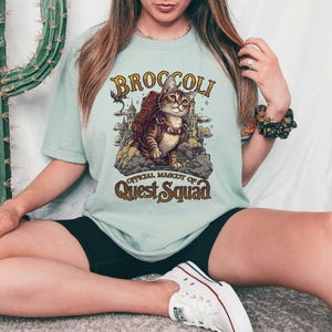 Pu&ograve; includere: Una maglietta blu chiaro con una grafica di un gatto che indossa uno zaino e il testo "Broccoli, mascotte ufficiale di Quest Squad".