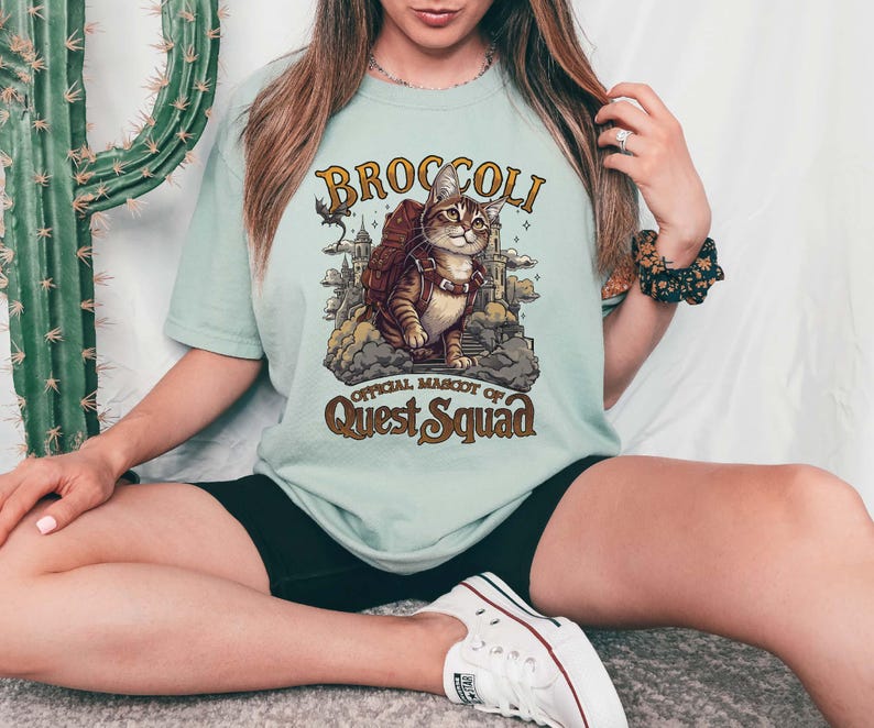 Pu&ograve; includere: Una maglietta blu chiaro con una grafica di un gatto che indossa uno zaino e il testo "Broccoli, mascotte ufficiale di Quest Squad".
