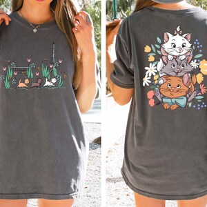 May include: A gray t-shirt with a cartoon image of three cats on the back. The cats are stacked on top of each other and surrounded by flowers. The front of the shirt has a cartoon image of three cats walking in a park.