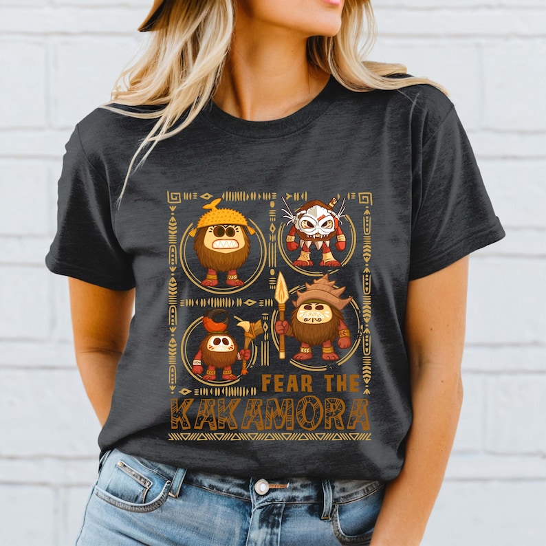 May include: Dark gray t-shirt featuring a graphic of four cartoonish figures with tribal designs. The figures are brown with various accessories and are set within decorative circles. The text "FEAR THE KAKAMORA" is printed in gold.
