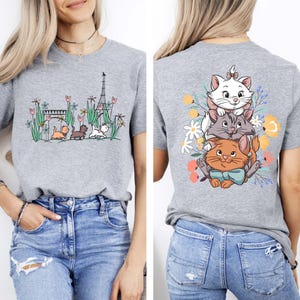 May include: Gray t-shirt with a graphic of three cats stacked on top of each other in front of flowers. The back of the shirt has a graphic of the Eiffel Tower, a bridge, and three cats walking in a line.