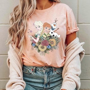 May include: A pink t-shirt with a graphic print of Elsa and Anna from Frozen, along with Olaf, surrounded by flowers.