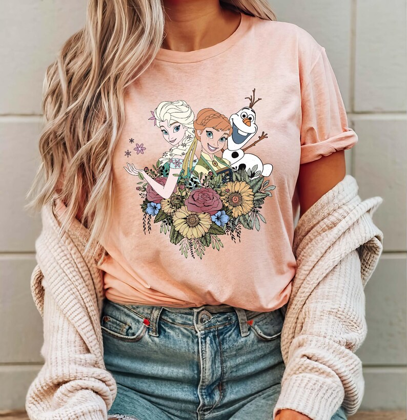 May include: A pink t-shirt with a graphic print of Elsa and Anna from Frozen, along with Olaf, surrounded by flowers.