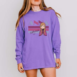 May include: A long-sleeved purple shirt with a cartoon graphic of a character with blonde braids and a purple cape. The shirt has the name "Anna" in a script font, with "ANNA" above it in a geometric font.