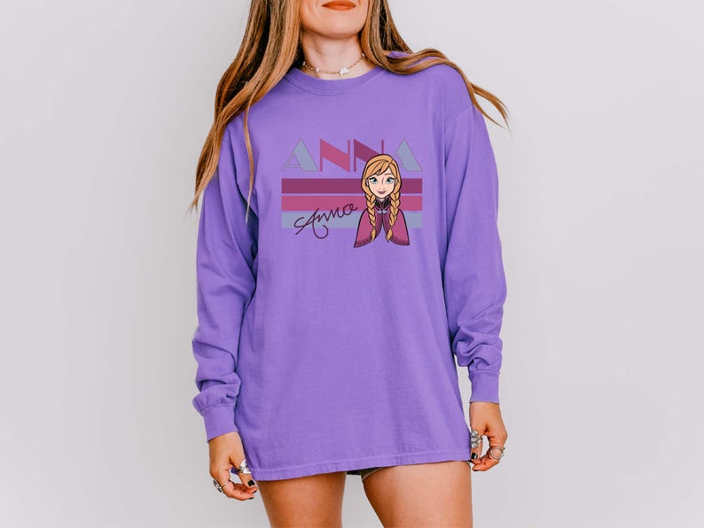 May include: A long-sleeved purple shirt with a cartoon graphic of a character with blonde braids and a purple cape. The shirt has the name "Anna" in a script font, with "ANNA" above it in a geometric font.