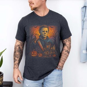 May include: Dark gray t-shirt featuring a Halloween-themed graphic. The design includes a figure holding a bloody knife, pumpkins, a haunted house, and bare trees under an orange sky. The shirt is short-sleeved.