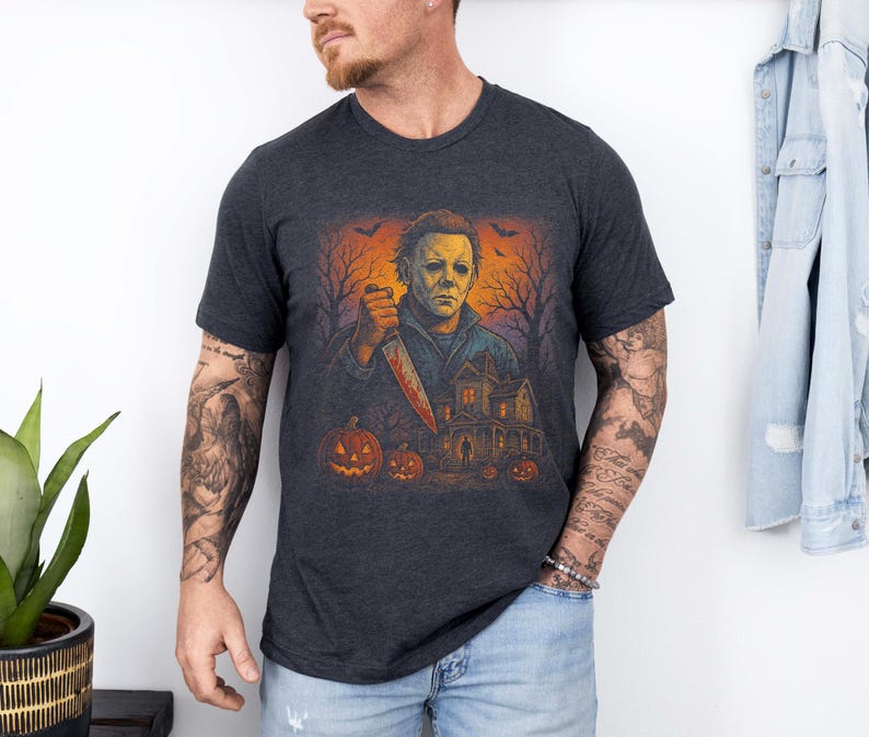 May include: Dark gray t-shirt featuring a Halloween-themed graphic. The design includes a figure holding a bloody knife, pumpkins, a haunted house, and bare trees under an orange sky. The shirt is short-sleeved.