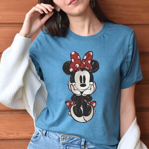 May include: A blue t-shirt with a cartoon image of Minnie Mouse wearing a red bow with white polka dots. Minnie Mouse is smiling and has her hands clasped in front of her.