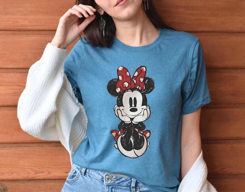 May include: A blue t-shirt with a cartoon image of Minnie Mouse wearing a red bow with white polka dots. Minnie Mouse is smiling and has her hands clasped in front of her.