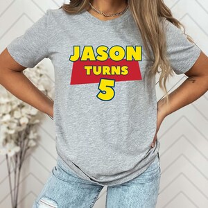 May include: A heather gray short-sleeve t-shirt featuring a graphic print with the text JASON TURNS 5. The word JASON is in yellow, the word TURNS is in yellow on a red trapezoid background, and the number 5 is in large yellow font.