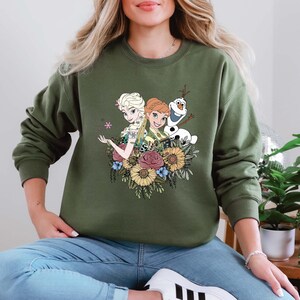 Pu&ograve; includere: Una felpa verde con un design floreale con i personaggi Elsa e Anna di Frozen, insieme a Olaf.