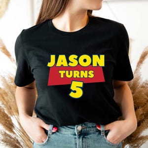May include: A black short-sleeved t-shirt featuring a graphic design with the text JASON TURNS 5. The word JASON is in yellow, the word TURNS is in yellow on a red trapezoid background, and the number 5 is in yellow with a blue outline.