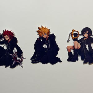 May include: Three anime-style character acrylic charms featuring figures in black hooded coats. The left character has spiky red hair and circular weapons, the center has spiky blonde hair, and the right has dark hair and a large key-shaped sword.
