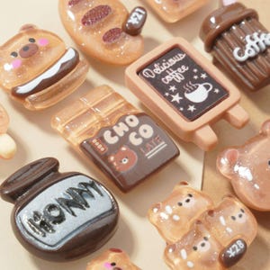 May include: A collection of miniature food-themed charms, including a bear sandwich, chocolate bar, coffee sign, and honey pot. The charms are in shades of brown, beige, and white, with some glitter accents. The sign reads "Delicious coffee".