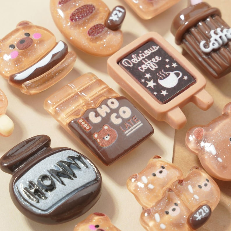 May include: A collection of miniature food-themed charms, including a bear sandwich, chocolate bar, coffee sign, and honey pot. The charms are in shades of brown, beige, and white, with some glitter accents. The sign reads "Delicious coffee".