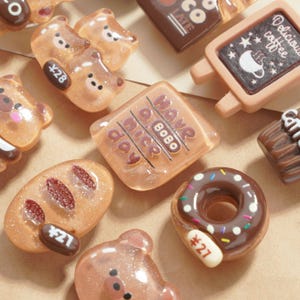 May include: A collection of miniature, glittery resin food charms in various shapes. Includes bear shapes, a donut with sprinkles, a bread loaf, and a sign that says "Delicious coffee". Some charms have price tags.