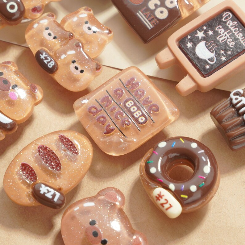 May include: A collection of miniature, glittery resin food charms in various shapes. Includes bear shapes, a donut with sprinkles, a bread loaf, and a sign that says "Delicious coffee". Some charms have price tags.