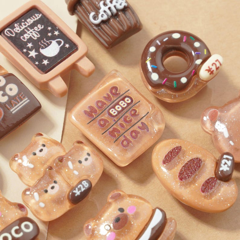 May include: A collection of miniature food-themed charms, including a donut with sprinkles, a bear-shaped candy, and a sign that says "Delicious coffee." Other charms include a chocolate bar and a piece of bread. The charms have glitter accents.