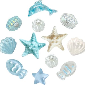 May include: A collection of ocean-themed embellishments, including a dolphin, starfish, shells, and fish. The pieces are in shades of blue, white, and beige, with some featuring glitter accents. These decorative elements are ideal for craft projects.