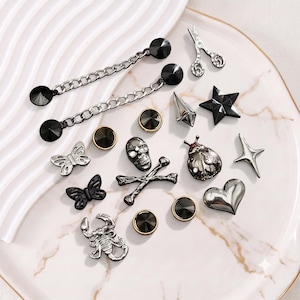 May include: A collection of decorative metal pins and buttons in various shapes including a skull and crossbones, butterflies, a scorpion, a ladybug, a star, a heart, scissors, and circular studs, all featuring a polished dark silver finish.