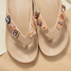 May include: Tan flip-flops adorned with various food-themed charms, including a coffee cup, bear, bread, and donut. The flip-flops have a textured footbed and a thong strap. The charms are in shades of brown, beige, and white.