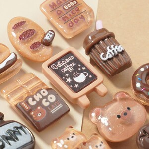 May include: A collection of miniature food-themed charms, including a chocolate bar, a coffee cup sign, a bear, a donut, and a coffee drink. The charms are in shades of brown, tan, and pink, with glitter accents. The chocolate bar has the text "CHO CO LATE".