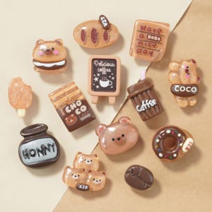May include: A collection of miniature food and bear-themed charms in shades of brown and beige. Includes a bear sandwich, a loaf of bread, a coffee cup, a chocolate bar, a donut, and a coffee bean. Some charms have text like "HONNY" and "CHO CO LATE".