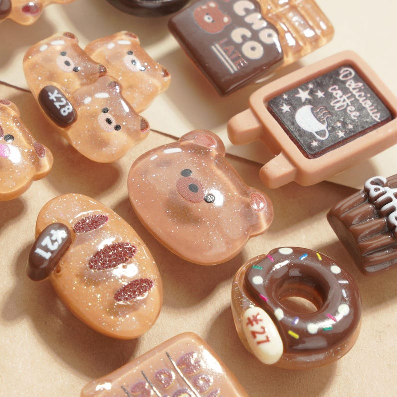 May include: A collection of miniature food-themed charms in shades of brown and tan. The charms include bear shapes, a donut, a loaf of bread, and a sign that says "Delicious coffee". Some charms have price tags.