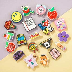 May include: A collection of colorful silicone shoe charms featuring school and teacher-themed designs, including a yellow school bus, a red apple with text, a stack of books, a pencil, a backpack, a clock, a smiley face, and a pig character.
