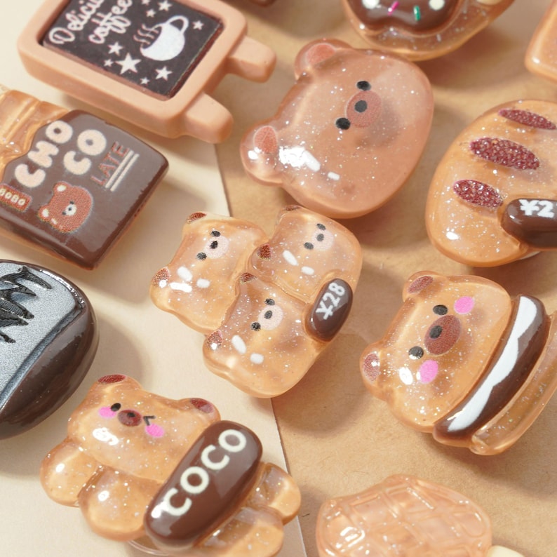 May include: A collection of various brown and tan resin charms, including bear shapes, a chocolate bar, and a coffee sign. Some charms have glitter accents and text, such as "COCO" and "Delicious coffee".