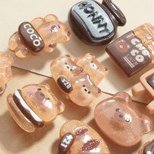 May include: A collection of various bear-shaped resin charms in shades of brown and tan, some with glitter accents. The charms include a bear with a "COCO" label, a honey jar, and a "Choco Latte" bar.