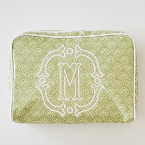 May include: A rectangular sage green zippered pouch featuring a repeating white fan-shaped pattern and a white piped edge. The center of the pouch displays a large, white embroidered monogram letter M enclosed within an ornate, decorative frame.