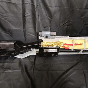 May include: A futuristic-looking, custom-built device with a complex design. It features a gray scope, black and clear plastic components, and yellow and gold internal elements. Red wires are visible, adding a pop of color to the overall aesthetic.