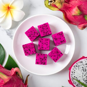 May include: A white plate with six pink dragon fruit jelly cubes. The plate is surrounded by dragon fruit, a green leaf, and a white flower.
