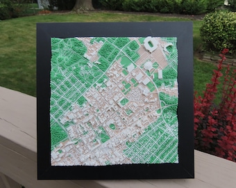 May include: A framed 3D topographic map featuring a city layout with white building structures and green textured areas representing parks and vegetation. The map is encased in a black square frame, displaying a detailed aerial view of urban streets.
