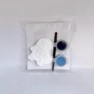 May include: A craft kit in a clear plastic bag containing a white turtle-shaped plaster figure, a thin black paintbrush, and two small circular containers of paint, one filled with dark blue paint and the other with light blue paint.