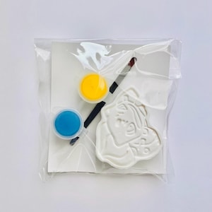 May include: A craft kit sealed in a clear plastic bag containing a white molded figure with a cartoon character design, a small paintbrush with a black handle, and two circular paint pots filled with yellow and blue paint on a white card.